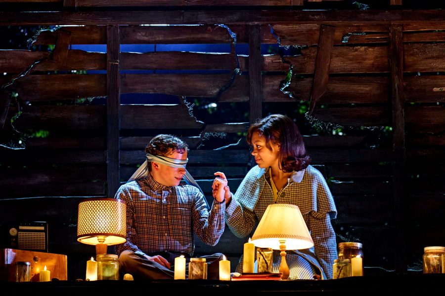 On stage photo. A woman (right) holds the hand and smiles at a boy (left) with a blindfold covering his eyes. There is an old wooden wall behind and lamps & candles in the foreground.