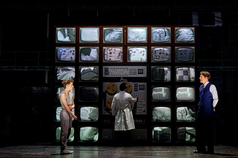 A grid of TV screens surrounding a panel of buttons & dials are in the background. In the foreground, a man in a blue suit & waistcoat (right) looks towards a young boy in a grey tracksuit (left).