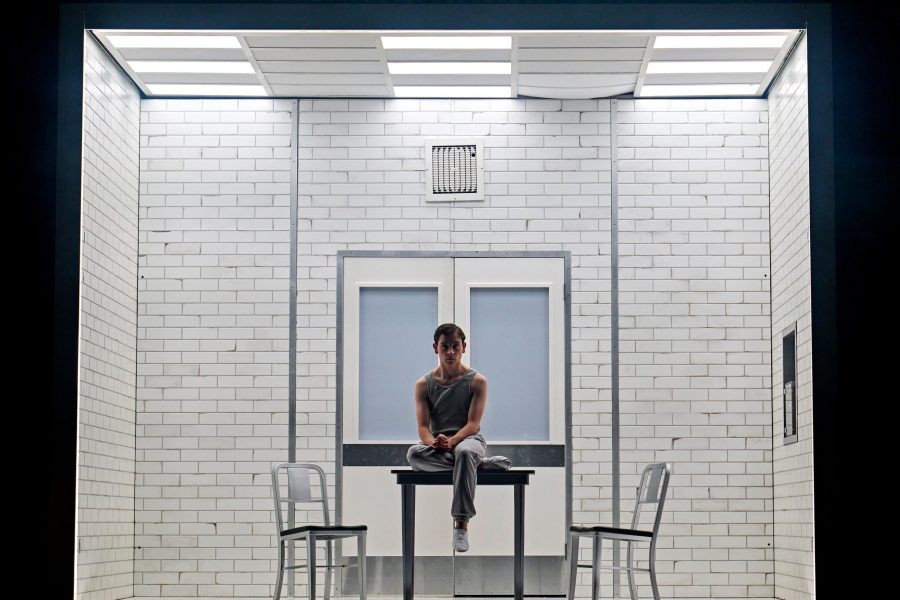 A lone boy wearing a grey tracksuit sits on a metal table with two empty chairs on either side. He is inside a white tiled room with air vents above and a double door behind.