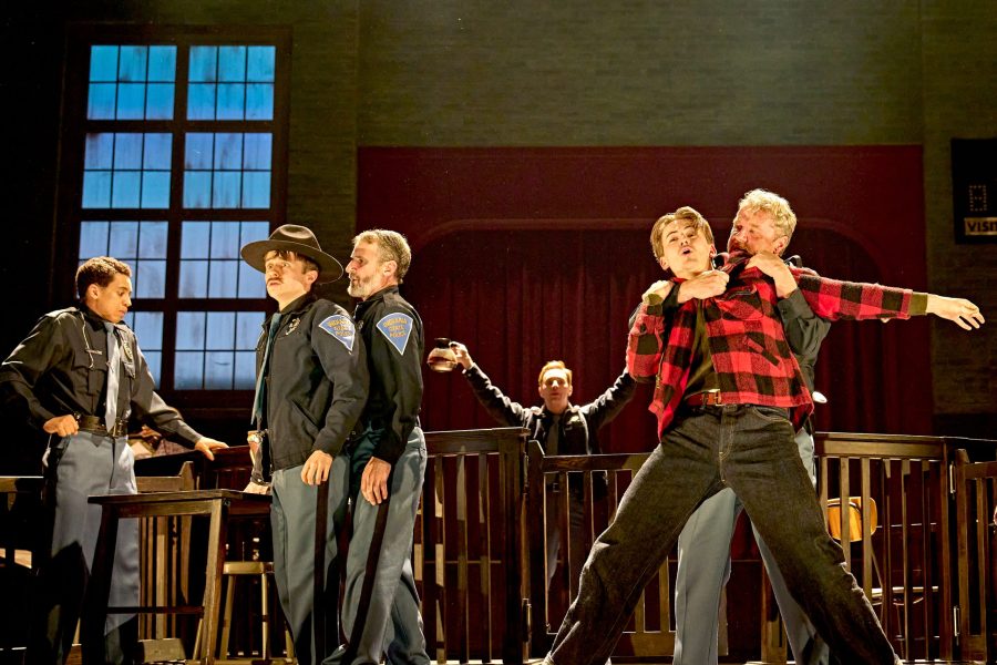 On-stage photo. Right: A burly man grapples with a younger teen in a red checked shirt. Left: A group of local policeman look worried. One man stands in the background brandishing a pot of coffee.